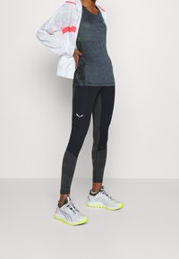 Sportovní outfit sestávající z bílé bundy s růžovými akcenty, tmavě modrého tílka, legín s černým a šedým vzorem a šedých bot s neony.