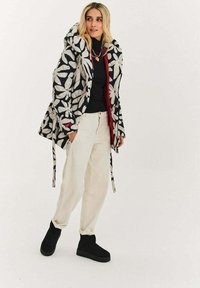 Černobílý kabát s květinovým vzorem, s kapucí, zapínáním na zip a šňůrkami, nošený přes černý top a krémové kalhoty, doplněný černými botami.