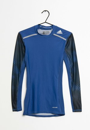 Langærmet blå Adidas atletisk kompressionsshirt med sorte mønstrede ærmer hængende på en sort bøjle mod en hvid baggrund.