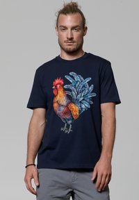 Marineblaues T-Shirt mit einem bunten Hahn-Graphic. Kurze Ärmel, Rundhalsausschnitt, aus Baumwolle und mit detaillierten Federakzenten in verschiedenen Farbtönen.
