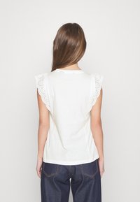 White sleeveless top with scalloped cotton eyelet detailing at shoulders, soft texture, and relaxed fit. Paired with dark denim bottoms.