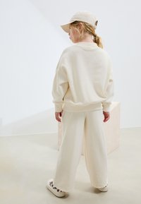 Sweat-shirt en coton couleur crème et pantalon large assorti, associés à une casquette beige et des baskets à motifs, vus de dos.