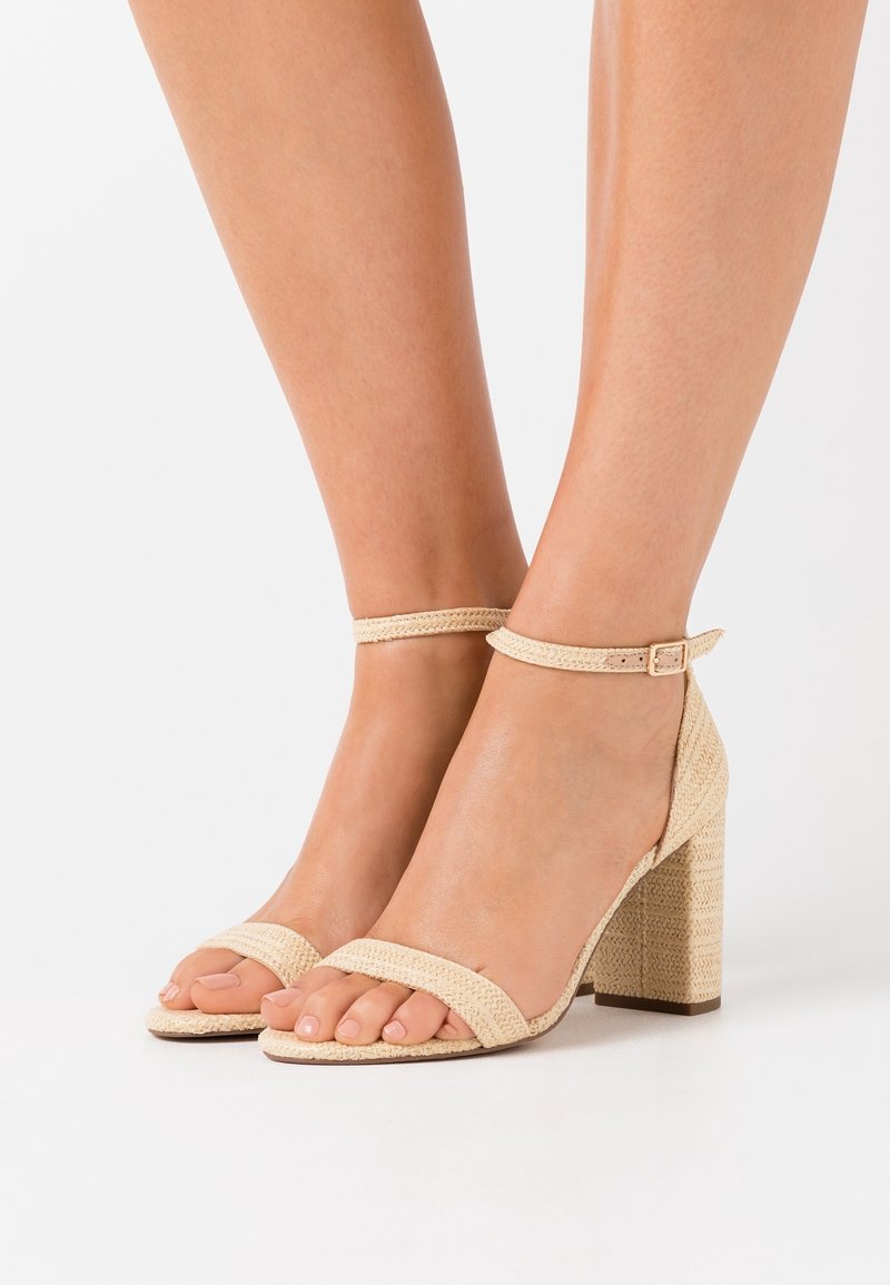 Pieds portant des sandales beige tissées à talons blocs avec des brides autour de la cheville, posés sur un fond blanc uni.