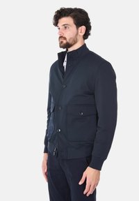 Giacca navy con colletto alto, bottoni frontali, due tasche sul petto e polsini elasticizzati. Tessuto liscio e vestibilità sartoriale, adatta per essere indossata in sovrapposizione.