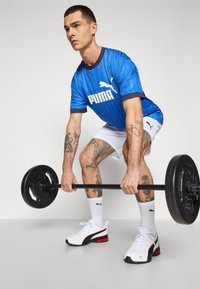 Blå stribet atletisk skjorte med hvide grafikker, hvide shorts og hvide sneakers med sorte og røde accenter, som holder en sort vægtstang.