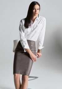 Witte button-up shirt met lange mouwen, gecombineerd met een nauwsluitende bruine leren rok met zijnaad en een gladde textuur. Gouden armbanden accentueren de look.