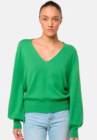 Un pull en tricot vert avec un col en V, de longues manches bouffantes et un ourlet côtelé. Texture douce avec une coupe décontractée. Porté avec un jean bleu clair.