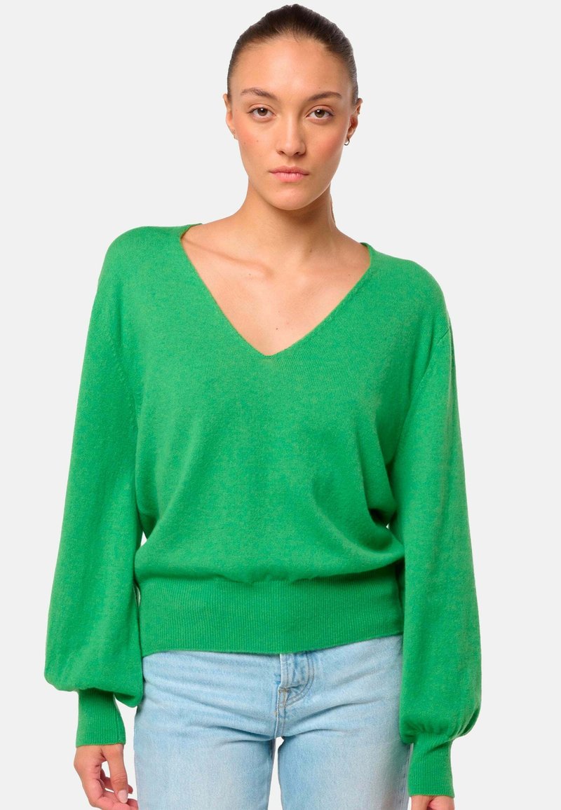 Un pull en tricot vert avec un col en V, de longues manches bouffantes et un ourlet côtelé. Texture douce avec une coupe décontractée. Porté avec un jean bleu clair.
