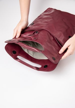Bordeauxleren tas met een ritssluiting, drukknoopsluitingen en stofvoering. Heeft een handvat aan de bovenkant en een ruime opening voor gemakkelijke toegang.