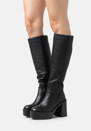 Botas altas de cuero negro con acabado texturizado, un tacón grueso y una cremallera lateral para facilitar su uso, con un diseño ligeramente arrugado.