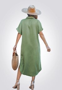 Olijfgroene, kortgebouwde jurk met korte mouwen, een kraag, zijsplitsen en een afgeronde zoom. Geweven handtas en lichte blokhak sandalen.