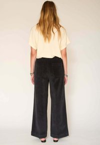 Pantalons en velours côtelé noirs à jambe large avec deux poches arrière, associés à une chemise à manches courtes beige clair, présentés de dos.