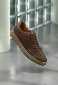 Zapatilla de ante marrón con un acabado texturizado, suela de goma crema y acentos beige. Presenta cordones y un logo sutil en el lateral.