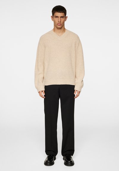 Beige bordázott V nyakú pulóver, oversize fazon, fekete egyenes szabású nadrággal és fekete fűzős cipővel viselve. Egyszerű dizájn, texturált anyag.