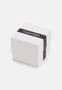 Valge kartongkarp musta pabeririba peale. Riba peal on valge tekstiga 'Calvin Klein', luues kontrasti ja brändingu.