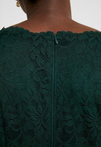 Grön spets tyg med blommönster, med en dragkedja bak. Texturen  är intrikat, med lager av detaljer och en scalloped-halsringning.