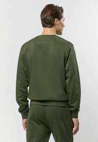 Tumši zaļš krūšturis ar garām piedurknēm, izgatavots no mīksta auduma, ar ribotiem piedurkņu galiem. Aizmugures skats parāda gludu tekstūru bez rakstiem.