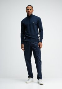 Navy gebreide pullover met een kraag met rits, gecombineerd met navy broek en witte sneakers. Beschikt over een subtiel textuurpatroon en logo.