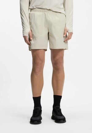 Mann trägt beige Shorts, ein passendes langärmeliges Hemd, schwarze Socken und schwarze Sportschuhe und steht vor einem schlichten weißen Hintergrund.