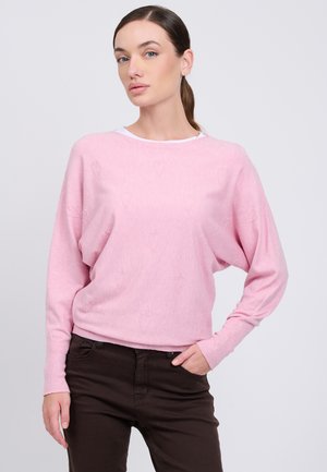 Femme aux cheveux bruns attachés en queue de cheval, portant un pull rose clair avec de subtils motifs en forme de cœur et un pantalon marron foncé, debout devant un fond uni.