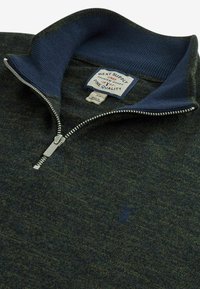 Tmavě zelený svetr s čtvrtým zipem, s žebrovaným námořnicky modrým límcem, zapínáním na zip a jemnou texturou; má logo na prsou a štítek s pokyny na péči uvnitř.