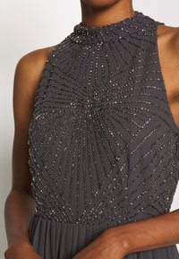 Grå ärmlös klänning med hög halsringning. Har pärlbroderi i en solstrålemönster över bystområdet. Plisserad kjol nedanför.