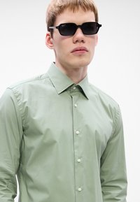 Jovem usando óculos de sol retangulares pretos e uma camisa de botão verde-claro diante de um fundo branco liso.