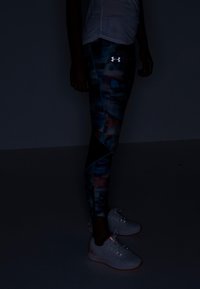 Kuvioitu leggings tummissa väreissä, joissa on sinisiä ja vaaleanpunaisia yksityiskohtia, yhdistettynä valkoisiin verkkomateriaalisempiin juoksukenkihin. Leggingseissä on heijastava logo.