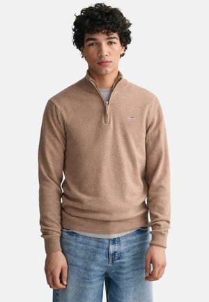 Maglione beige con zip a un quarto, realizzato in materiale morbido, con polsini e orlo a coste, presenta un piccolo logo sul petto. Indossato sopra una maglietta grigia.