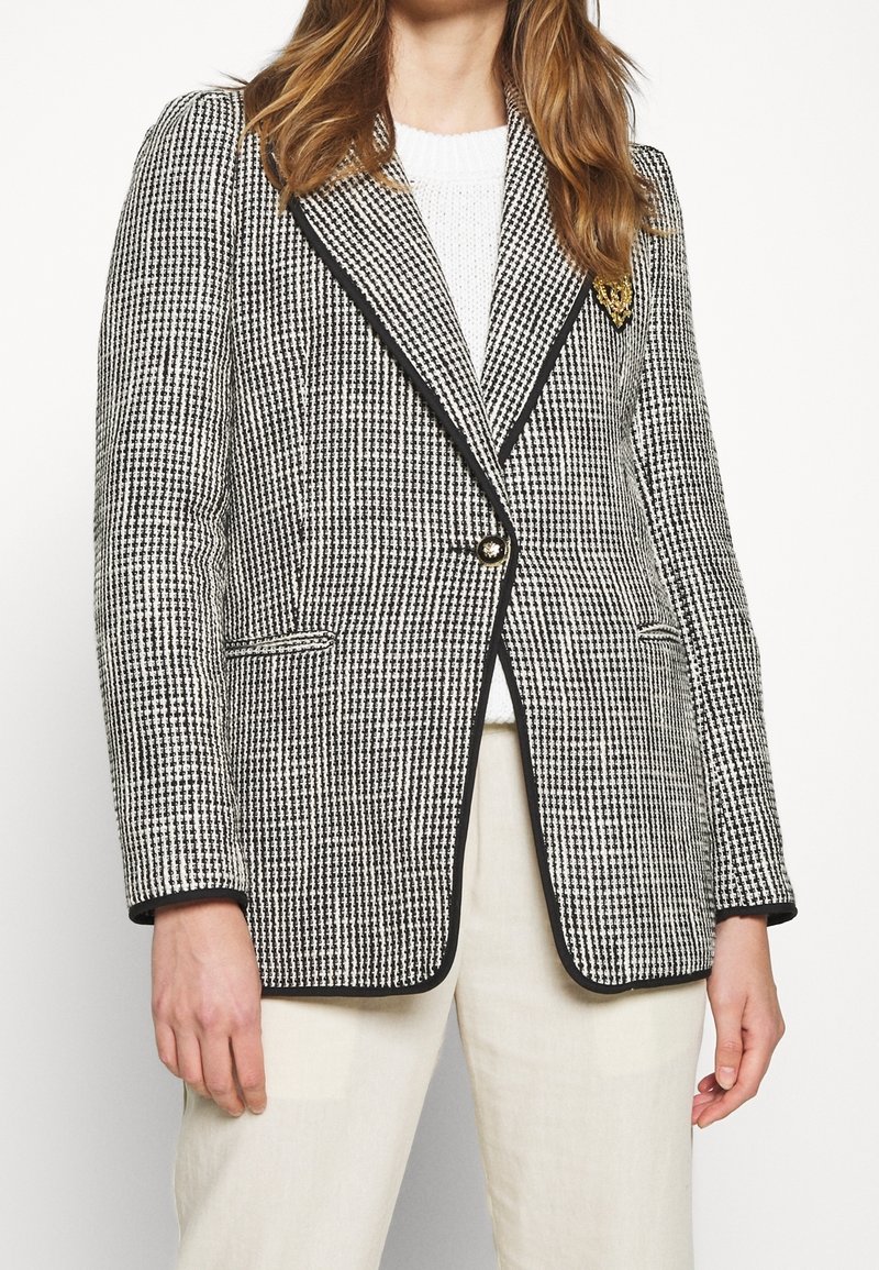 Femme portant un blazer pied-de-poule noir et blanc avec bordure noire sur un pull blanc et un pantalon crème, montrée des épaules jusqu'au milieu des cuisses.