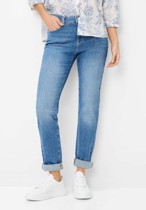 Frau trägt hellblaue Jeans mit Umschlag und eine Hand in der Tasche, kombiniert mit weißen Turnschuhen und einem Hemd mit Blumenmuster.