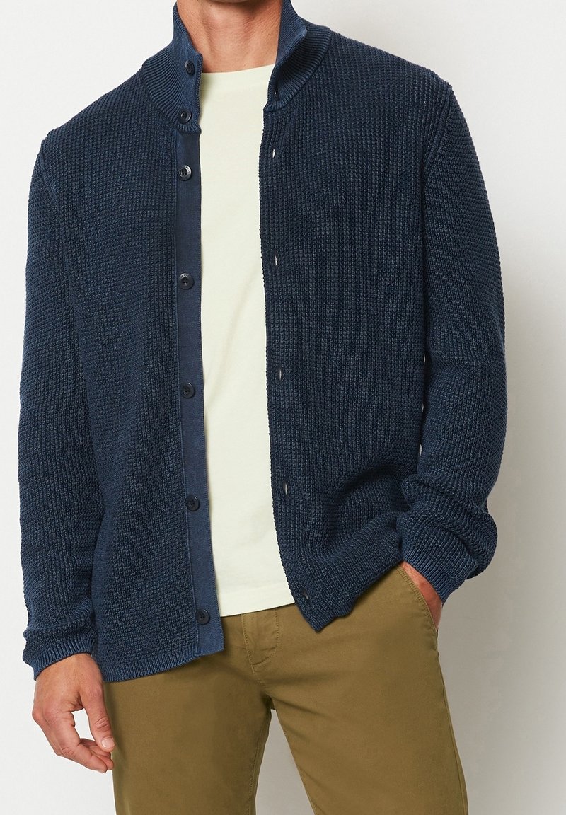 Marinblå stickad kofta med ett texturerat mönster, framknappar och hög krage. Bärs över en ljusfärgad skjorta och beige byxor.