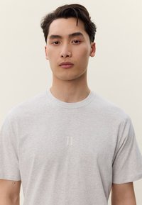 Mladý muž s krátkymi tmavými vlasmi, oblečený v svetlosivom tričku s malým symbolom dvoch zvislých čiar na hrudi, neutrálne pozadie.