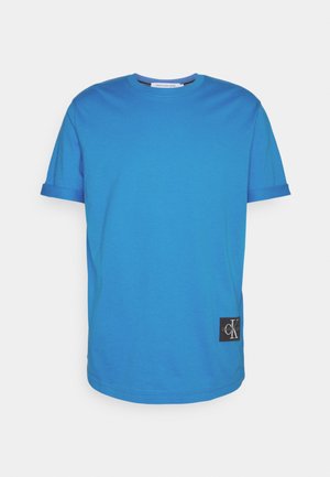 Niebieska bawełniana koszulka z krótkim rękawem, okrągłym dekoltem i czarną naszywką z logo Calvin Klein na dolnej lewej stronie. Gładka struktura.