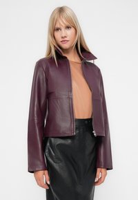 Veste en cuir bordeaux avec un col montant, fermeture éclair sur le devant et manches longues, associée à un haut nude et une jupe plissée noire.