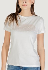 Camiseta de algodón blanca con cuello redondo y mangas cortas. Presenta un logo metálico de puntos "GUESS" en la parte frontal. Textura suave.