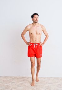 Sarkani peldkostīmi ar auklu, viegla materiāla, un mazs logotips priekšpusē. Models stāv basām kājām, rokas uz gurniem.