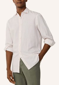 Mies, jolla on valkoinen pitkähihainen nappipaita, jossa ohuet pystysuorat vaaleanpunaiset raidat, sekä vihreät housut, hihat käärittynä ylös, toinen käsi taskussa.