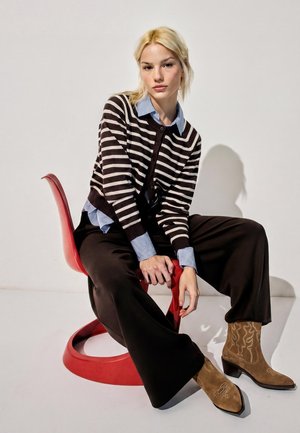 Giovane donna con capelli biondi seduta su una sedia moderna rossa, indossa un maglione a righe, una camicia blu con colletto, pantaloni neri larghi e stivali color tan ricamati.