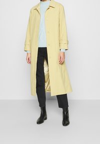 Ljusgul, lång kappa i bomullsblandning, med krage, knappstängning och uppvikta ärmar. Kombination med en ljusblå tröja och svarta jeans.