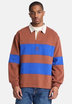 Mann trägt ein braun-blau gestreiftes Rugbyshirt mit langen Ärmeln, weißem Kragen und blauen Jeans, stehend vor einem einfarbigen Hintergrund.