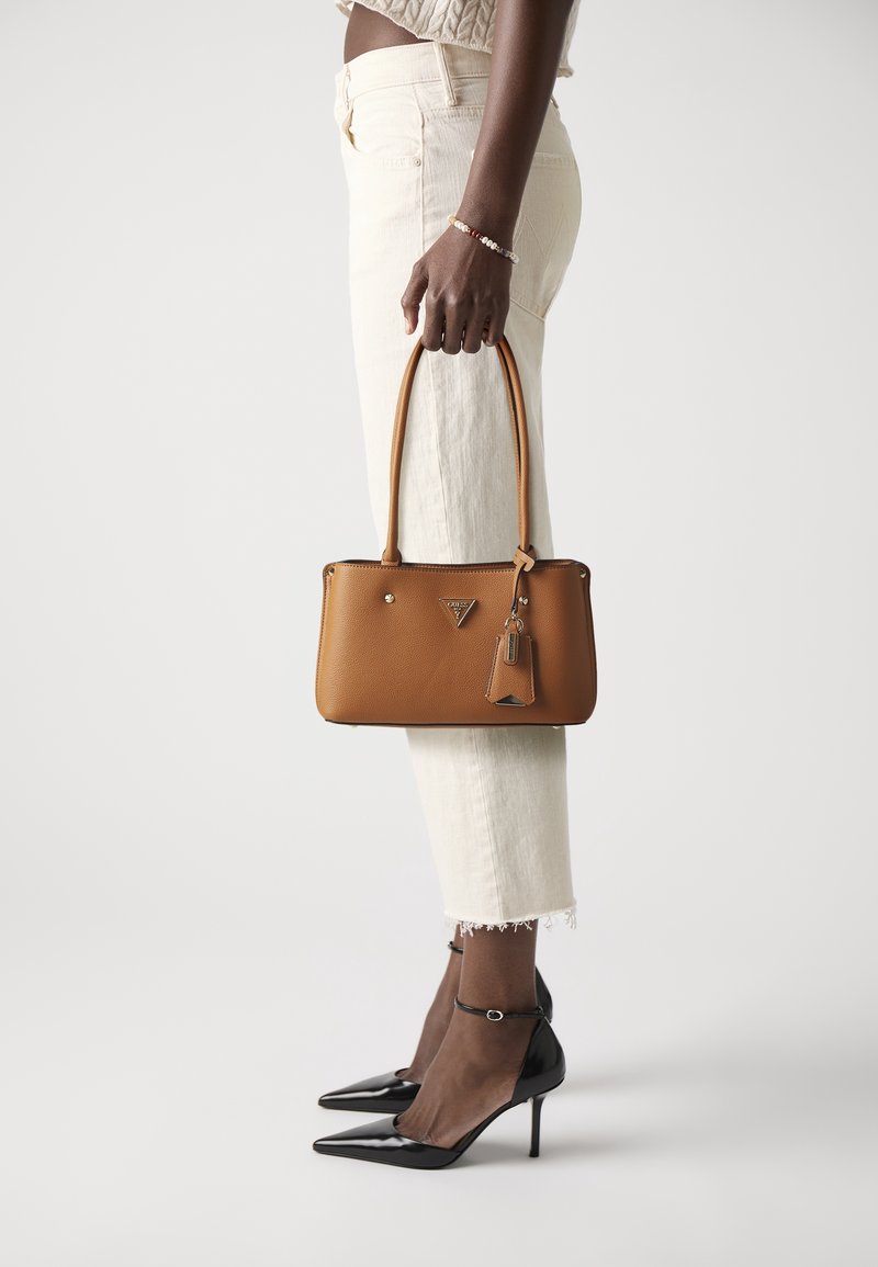 Femme portant un pantalon blanc court et des talons noirs tenant un sac à main en cuir marron par ses poignées à son côté.