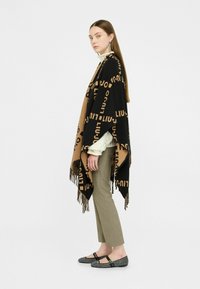 Gebreide poncho in zwart en kamel met gedurfde tekstpatronen, franjes aan de onderkant, gecombineerd met olijfgroene broek en zwarte pontjes met patroon.