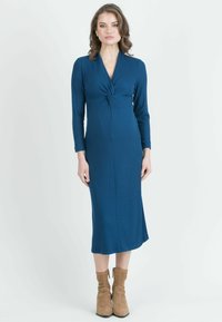 Donna che indossa un vestito blu a coste, lungo fino al ginocchio, con un dettaglio frontale attorcigliato e stivaletti beige in camoscio alla caviglia.