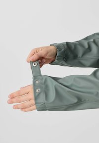 Ruky upravujú patentový gombík na manžete nepremokavej bundy tlmenozelenej farby na rukáve proti jednoliatemu pozadiu.