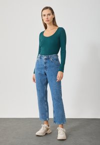 Zelený rebrovaný top s dlhým rukávom skombinovaný s vysokými modrými denimovými džínami. Model má na sebe svetlé tenisky. Jednoduché pozadie.