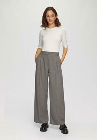 Haut blanc à manches courtes avec un col festonné, associé à un pantalon gris taille haute à jambes larges avec des plis et des poches latérales. Chaussures noires.