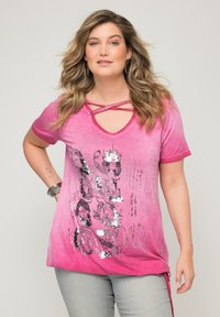 Vaaleanpunainen T-paita, jossa on V-kaula-aukko ja ristikkäinen yksityiskohta, metallinhohtoisella hopeisella graafisella painatuksella ja tekstillä, valmistettu pehmeästä kankaasta.