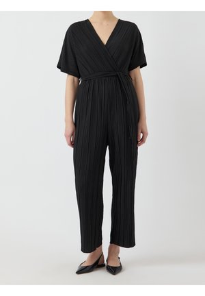 Frau trägt einen schwarzen, plissierten Jumpsuit mit kurzen Ärmeln, Wickel-V-Ausschnitt und gebundenem Gürtel in der Taille, kombiniert mit schwarzen spitzen Ballerinas.