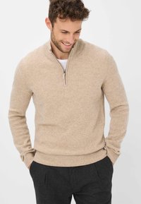 Beiger, gestrickter Pullover mit hohem Kragen und Viertelreißverschluss. Strukturierter Stoff, figurbetonte Silhouette und gerippte Bündchen. Über einem weißen Hemd getragen.